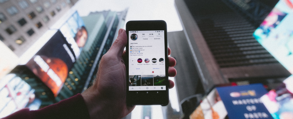 © Jakob Owens - Unsplash, Hand hält Smartphone vor Hochhäusern, auf dem Screen die Instagram App