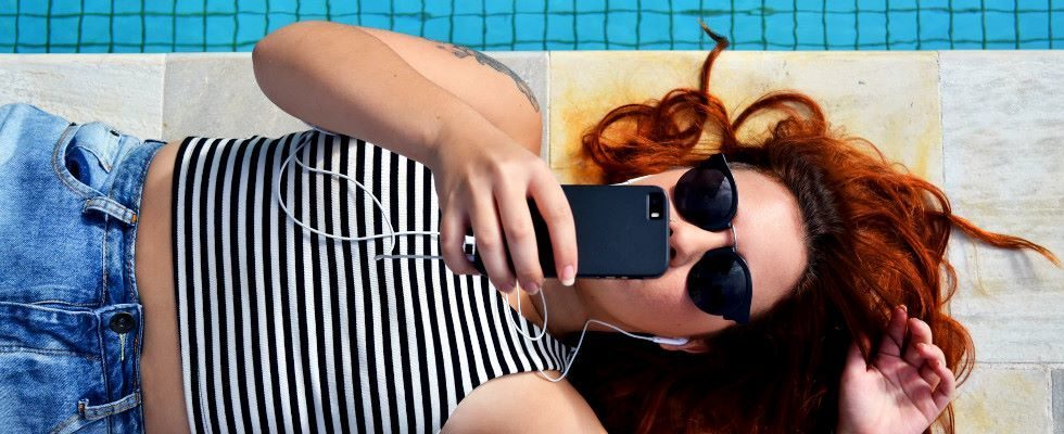 © Bruno Gomiero - Unsplash, Frau liegt mit Sonnenbrille und Smartphone und Kopfhörern neben einem Pool 