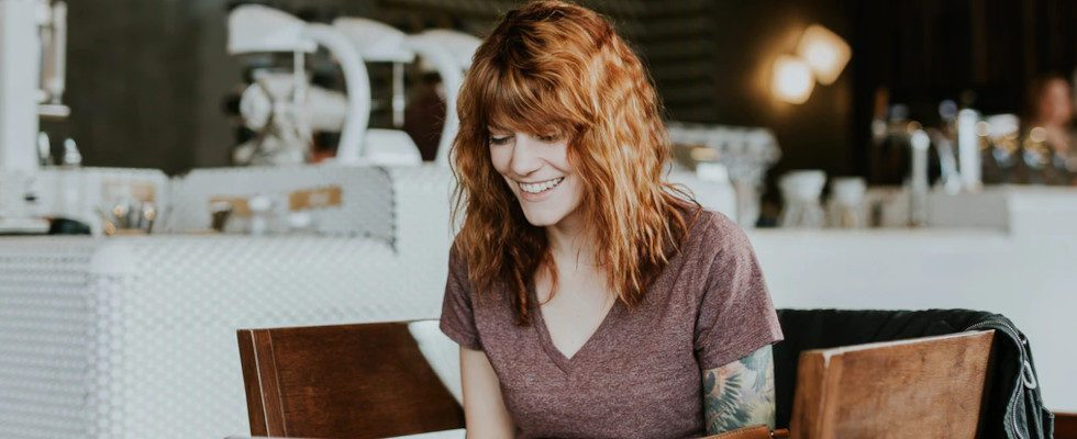 © Brooke Cagle - Unsplash, Frau mit rotem Haar sitzt lächeln am Tisch, Küche/Bar im Hintergrund
