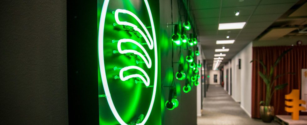 © Spotify via Canva, Spotify-Logo mit Leuchtröhren, grün leuchtend, an Wand, Büroflur mit Pflanze und Türen