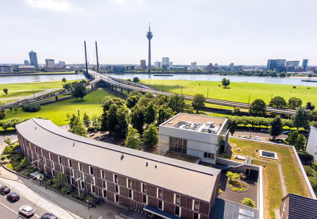 Mitten in Düsseldorf kannst du dir 2024 enorm viel Marketing Know-how sichern, © Rheinwerk, Gebäude vor Bäumen, Brücke und Skyline von Düsseldorf im Hintegrund