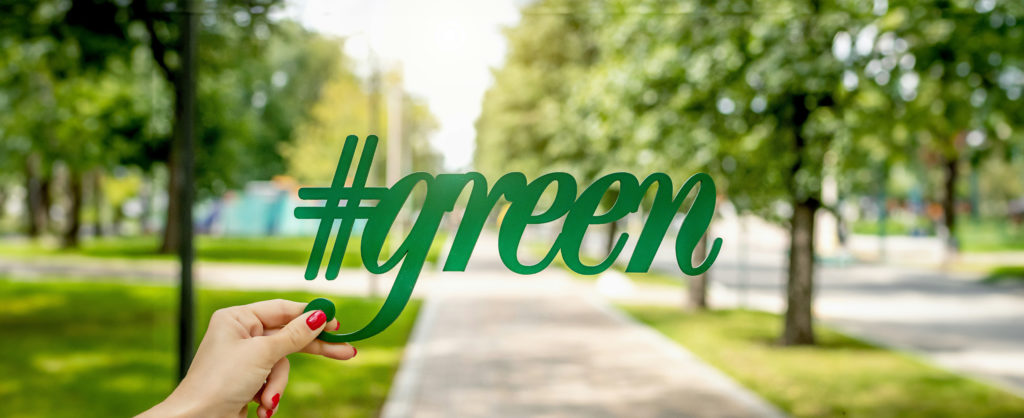 © Element C via Unsplash, Hand hält Hashtag mit Schriftzug green vor Weg mit Bäumen, verschwommen im Hintergrund