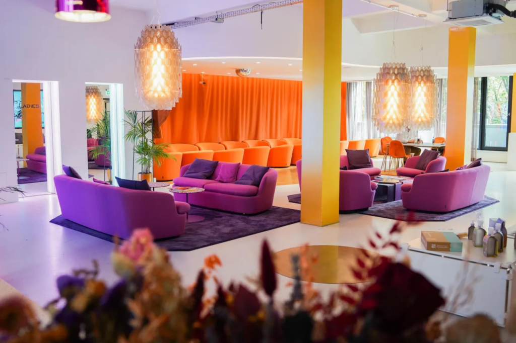 Raumansicht mit hellen Hängelampen, Sofas und Sesseln in orange und violett, heller Boden und helle Wände, Blumen und Tische sind zu sehen
