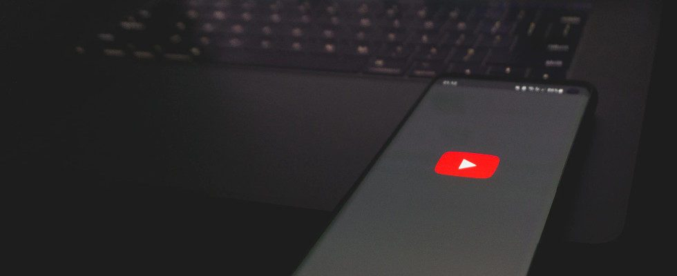 © Azamat E - Unsplash, dunkler Hintergrund, Handy mit YouTube-Logo auf Screen auf Tastatur von Laptop liegend