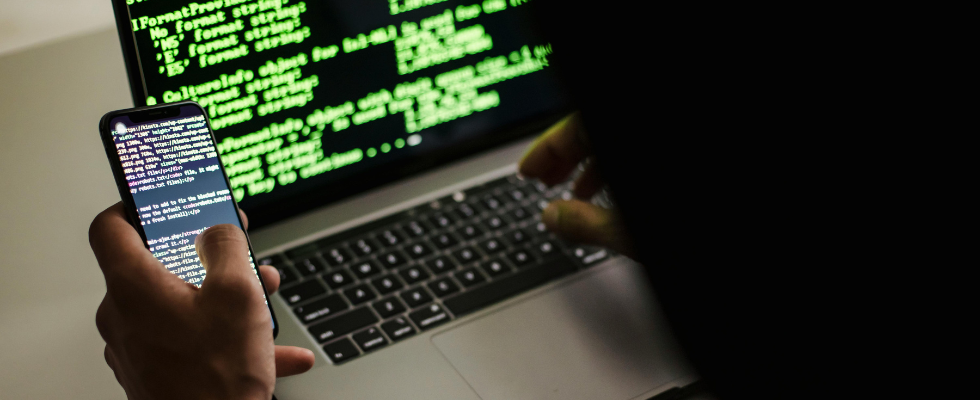 Ein Hacker sitzt vor einem Laptop und einem Smartphone