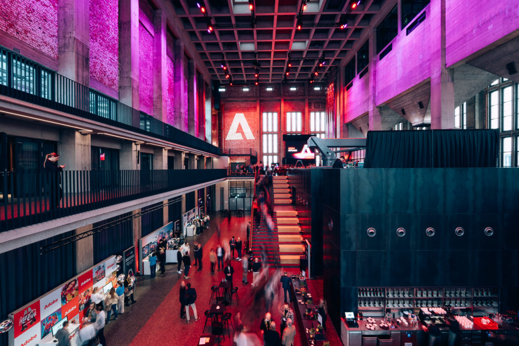 Blick in die Eventhalle des Bergson Kunstkraftwerks beim Adobe AI Forum München mit Demo-Ständen, Besucher:innen und Adobe Branding.
