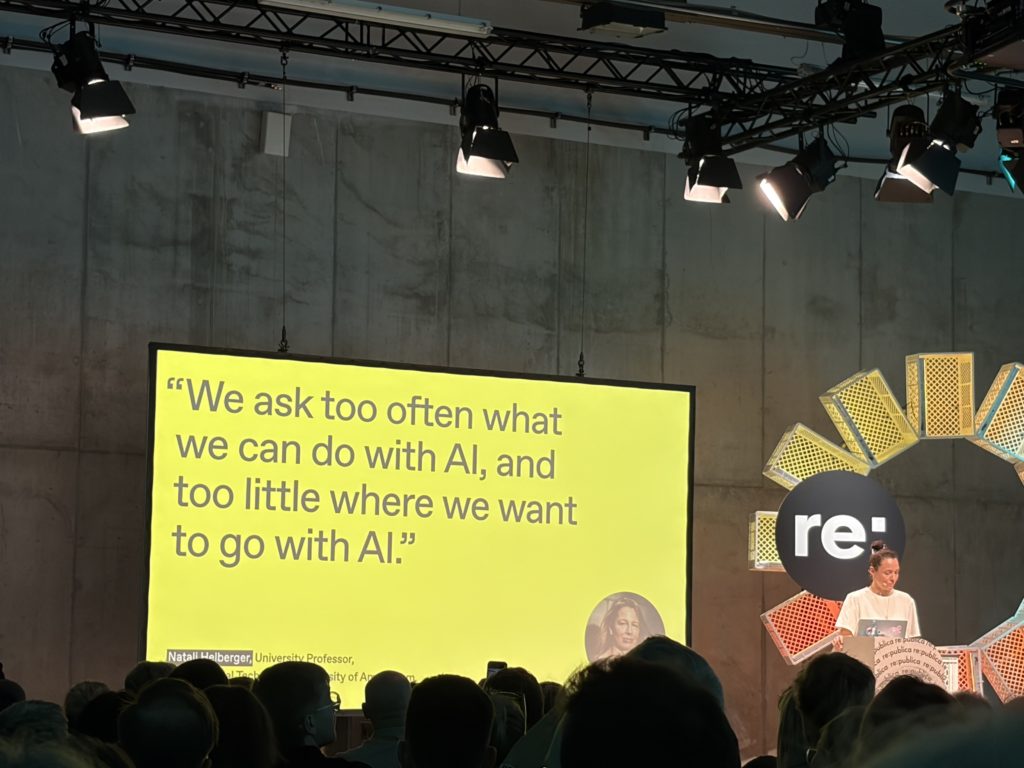 Eva-Maria Sommer spricht auf der re:publica 2026 auf einer Bühne vor Publikum. Hinter ihr ist eine große gelbe Präsentationsfolie mit einem englischen Zitat über den gesellschaftlichen Umgang mit KI und der Frage zu sehen, wohin sich KI entwickeln soll.