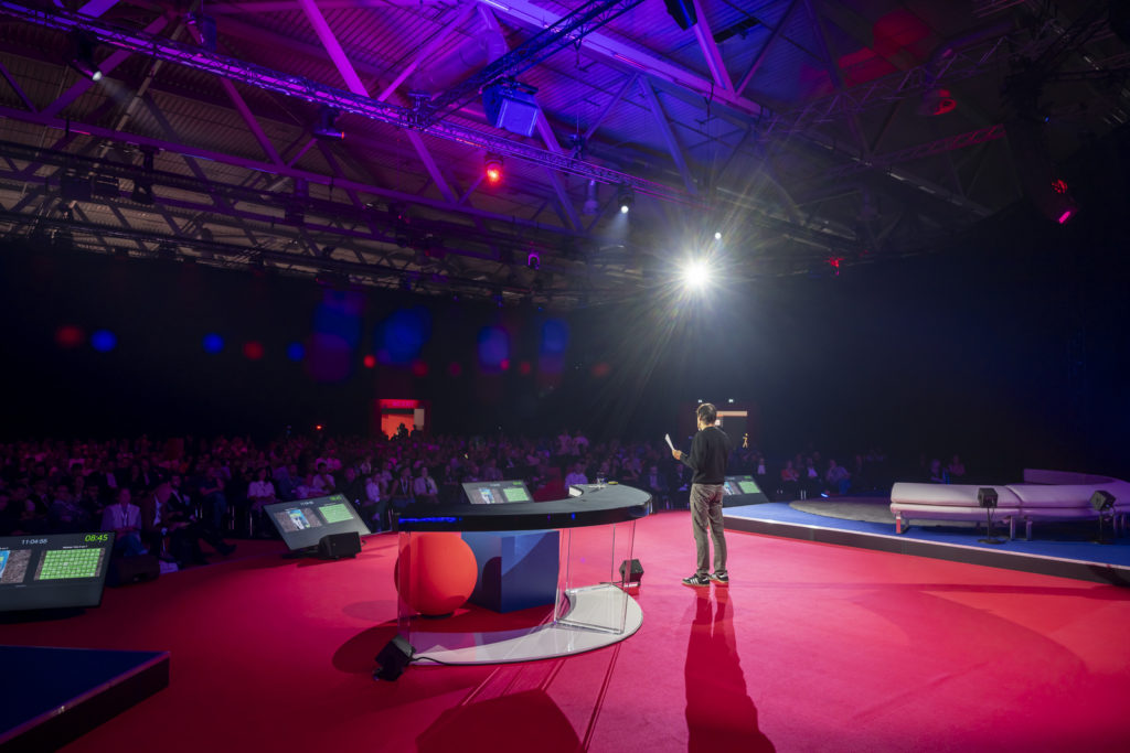 Die DMEXCO-Bühnen ziehen jedes Jahr Tausende Interessierte an, zu diversen Themen, © Koelnmesse, Person auf Bühne mit DMEXCO-Logo vor dem Tisch, dahinter ein großes Publikum, Lichtstrahl im Fokus