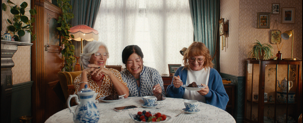 Gabi, Bärbel und Pham sitzen lachend bei Kaffee und Kuchen am Tisch. Die drei sind die Protagonistinnen der neuen ChatGPT Voice-Kampagne von OpenAI.